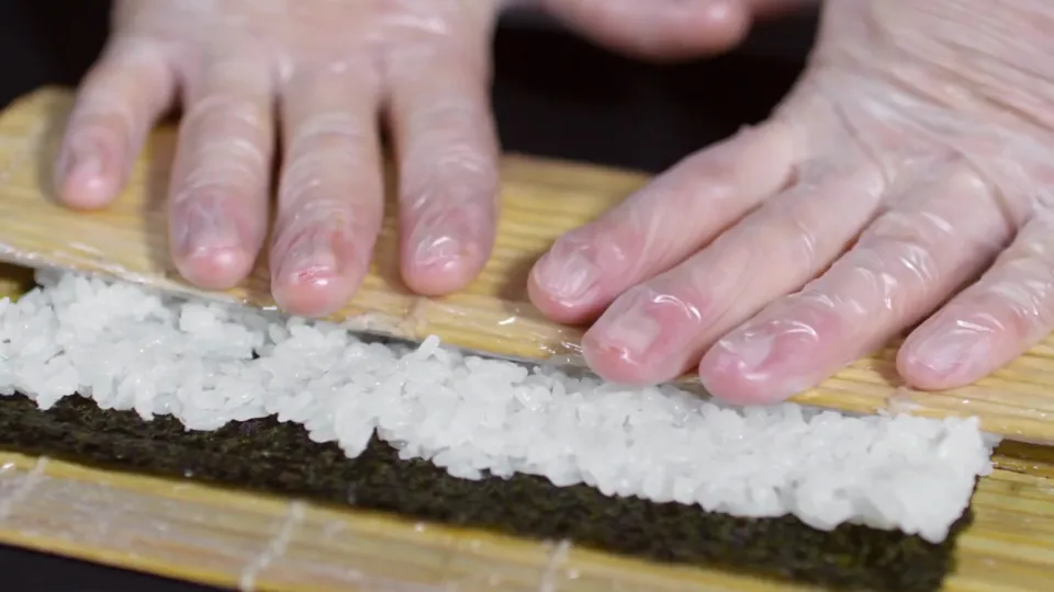 Mãos usando uma esteira para enrolar sushi sobre uma bancada